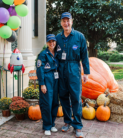 Ang Gobernador at Unang Ginang na nakasuot ng magkatugmang NASA astronaut jumpsuit ay magkasamang nag-pose sa labas sa panahon ng isang maligaya na kaganapan sa taglagas. Nakatayo sila sa harap ng isang display na nagtatampok ng malaking pumpkin, hay bale, at makukulay na lobo, na may maliit na laruang rocket sa malapit. Kasama sa setting ang isang brick walkway at isang klasikong white-columned na gusali, na napapalibutan ng mga napapanahong dekorasyon.