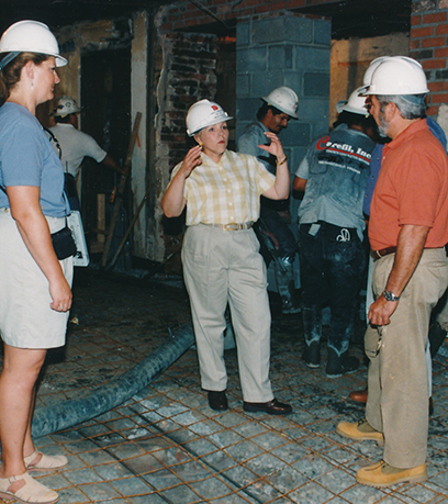 Nakatayo si First Lady Roxane Gilmore na nakasuot ng hard hat sa isang hindi pa tapos na istraktura at nakikipag-usap sa dalawa pang tao na nakasuot ng hard hat. Si Mrs. Gilmore ay nakasuot ng short sleeved button up shirt at khaki pants. Ang larawang ito ay kinuha sa pagitan ng 1998-2000.