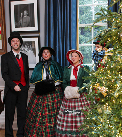 Tatlong performer na nakasuot ng period clothing ang nakatayo sa tabi ng isang nakasinding Christmas tree. Ang ilan ay nakabuka ang kanilang mga bibig, na nagpapahiwatig na sila ay kumakanta.