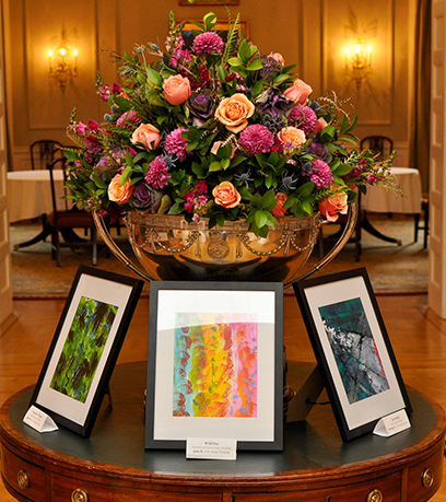 Tatlong painting sa itim na frame ang nakaupo sa isang round table na may pink na flower arrangement sa likod nila.