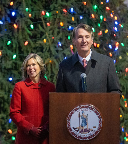 Si Governor Youngkin at First Lady Suzanne Youngkin ay nakatayo bilang isang podium sa harap ng isang malaking Christmas tree na may maraming kulay na mga ilaw.