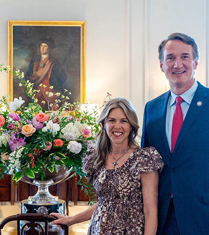 Ang Gobernador at Unang Ginang ay nag-pose sa harap ng floral arrangement sa Dining Room sa Historic Garden Day.