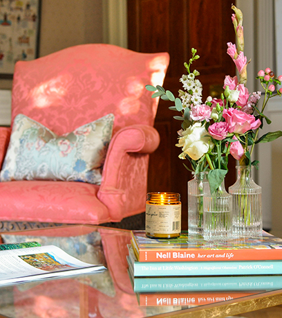 Isang coffee table na may tatlong bud vase na may mga bulaklak na nakaupo at isang kandila na nakapatong sa ibabaw ng isang stack ng mga libro. Ang isang coral chair na may pandekorasyon na unan ay nasa background.