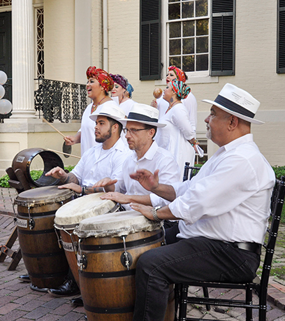 Ang Tradicion Dancy Company ay gumaganap sa labas ng Executive Mansion. Tatlong lalaki ang nakaupo na naglalaro ng drum sa harapan at isang grupo ng mga babae ang kumakanta sa likod nila.