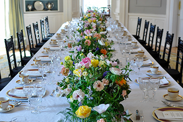 Dining table set na may puting table cloth at mga bulaklak na umaagos sa kahabaan ng mesa.